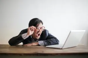 Modern Asian man in jacket and glasses looking at laptop and screaming with mouth wide opened on white background